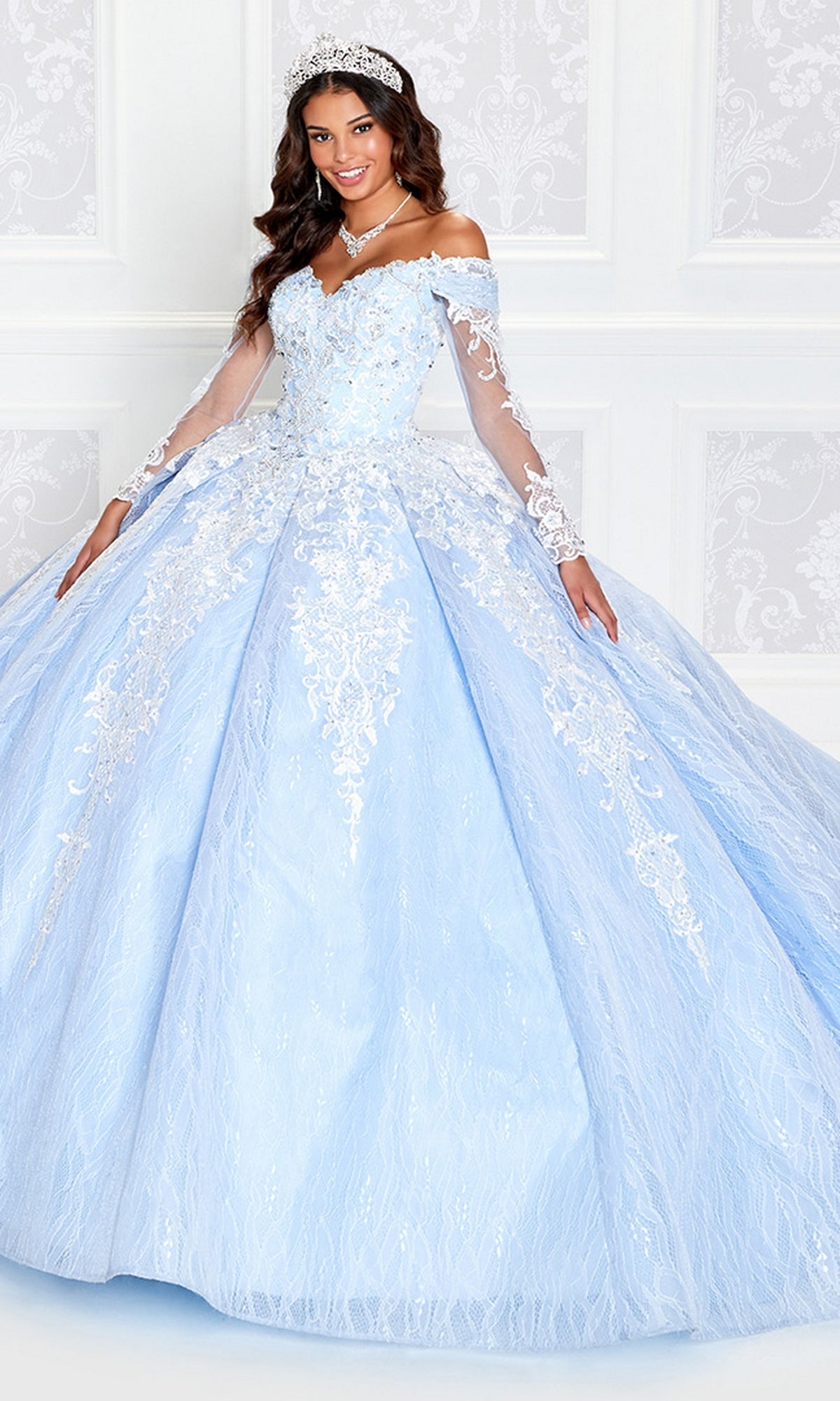 A woman in the Princesa Off-the-Shoulder Lace Quince Dress PR12267—light blue with lace, a lace-up corset back, and off-shoulder design—stands smiling by a white patterned wall, accessorized with a tiara and necklace.